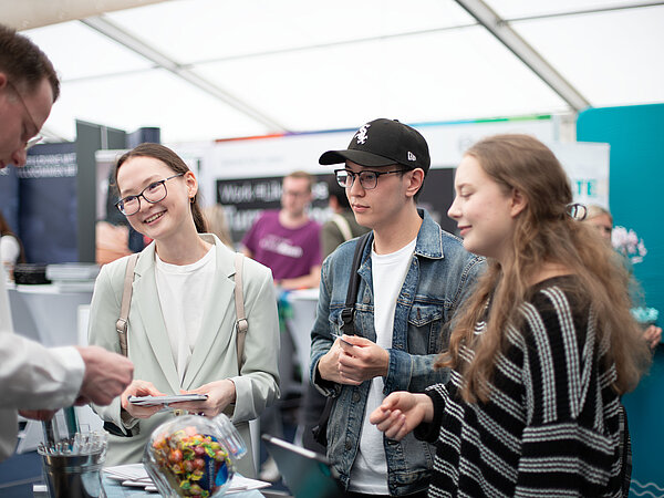 <img src="bild.jpeg"       alt="Drei junge Besucher im Gespräch mit einem Aussteller an einem Messestand, mit Infomaterial, Laptop und Bonbonbehälter auf dem Tisch.