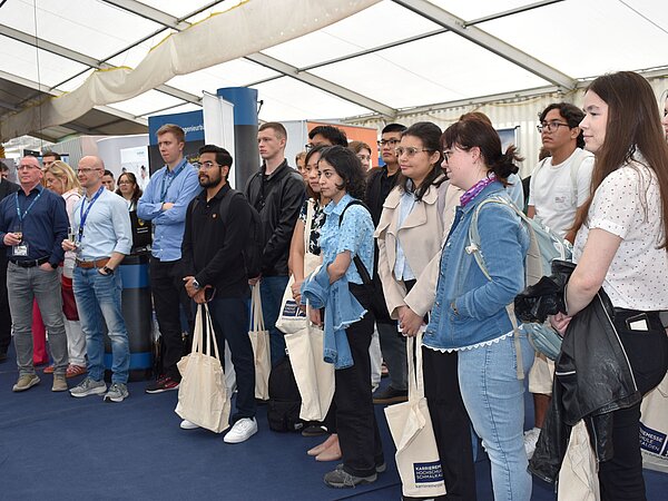 Besucher stehen in einem Messezelt und hören aufmerksam zu, umgeben von Informationsständen und Bannern. Viele tragen beigefarbene Stofftaschen mit einem Logo.