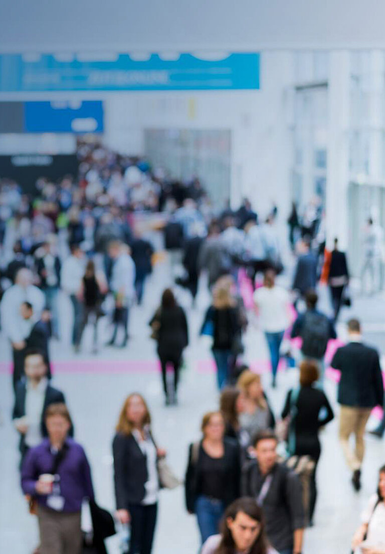 Visitors stream through a large exhibition hall with blue lighting while attending an event.