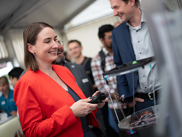 Frau im roten Blazer interagiert mit einem technischen Gerät auf einer Messe, umgeben von Besuchern und einem Mann im blauen Anzug.