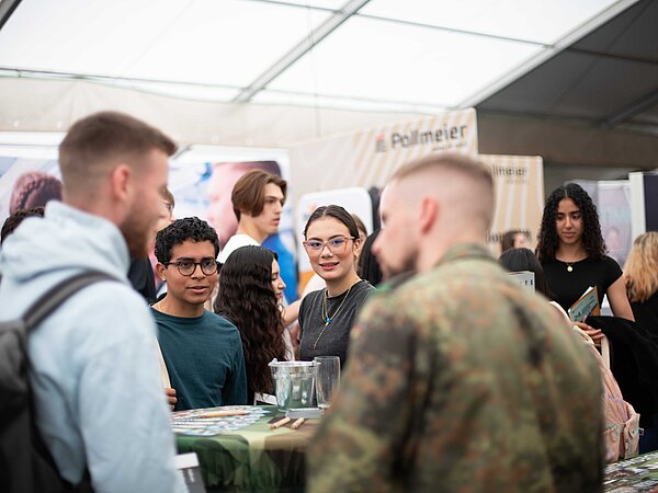 Besucherinnen und Besucher im Gespräch an verschiedenen Messeständen in einem großen Zelt. Eine Person in Tarnuniform steht im Vordergrund, umgeben von Infomaterial und Broschüren.