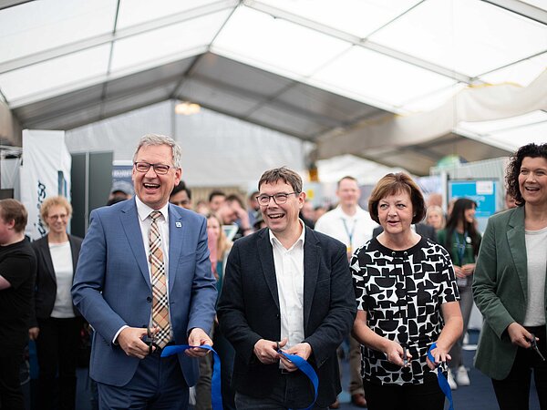 Vier Personen durchschneiden feierlich blaue Bänder bei einer Eröffnungszeremonie in einem Messezelt, umgeben von einer Menschenmenge und Informationsständen.