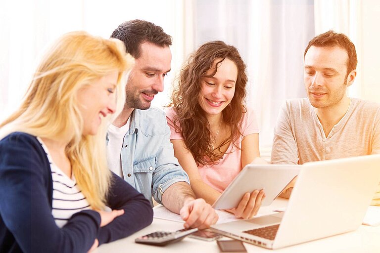 A working group smilingly discusses information on a tablet and laptop in a bright room.