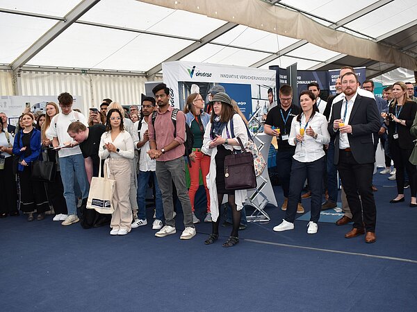 <img src="bild.jpeg"       alt="Menschen mit Namensschildern im Gespräch auf einer Messe mit blauer Teppichfläche, im Hintergrund ein Banner mit der Aufschrift 'enviss – Innovative Verschmutzungsbekämpfung'.