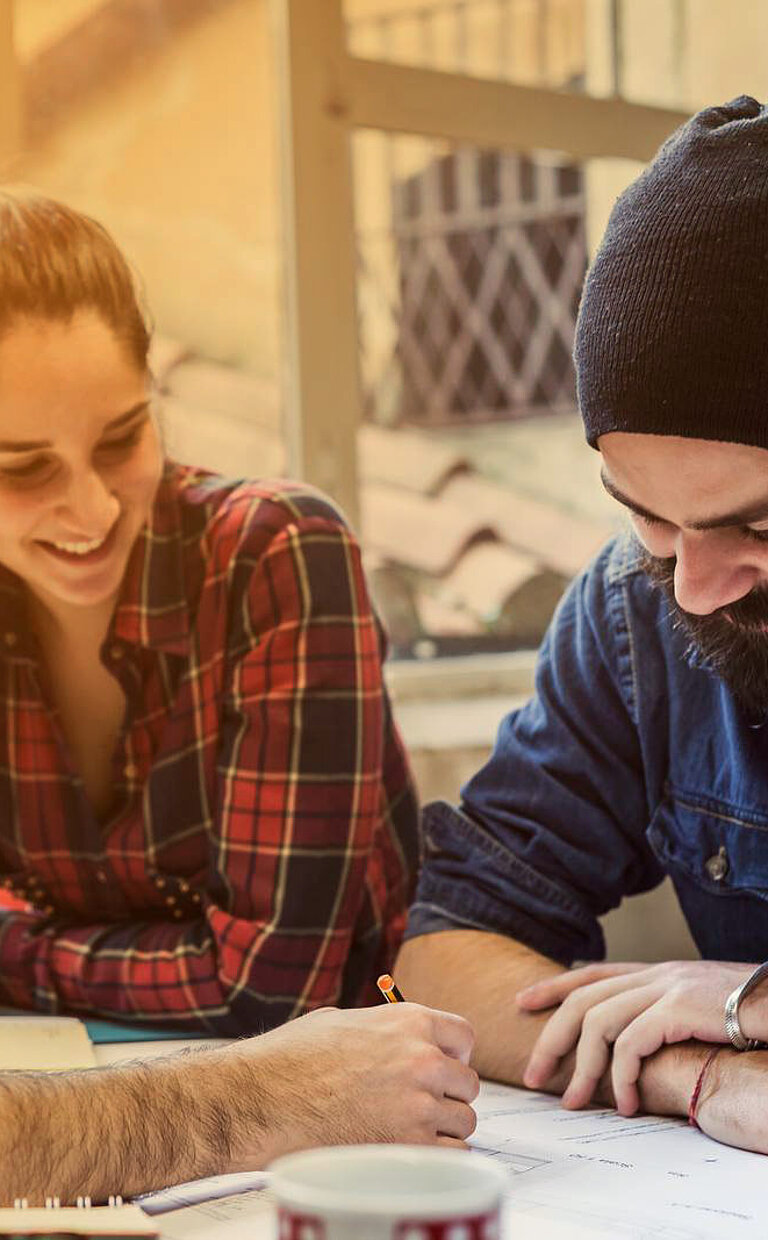 Three people sit together at a table, working intently on a project or task. One person is writing, while the others are attentively observing and discussing. The scene takes place in a bright interior with natural light, creating a relaxed and focused atmosphere.