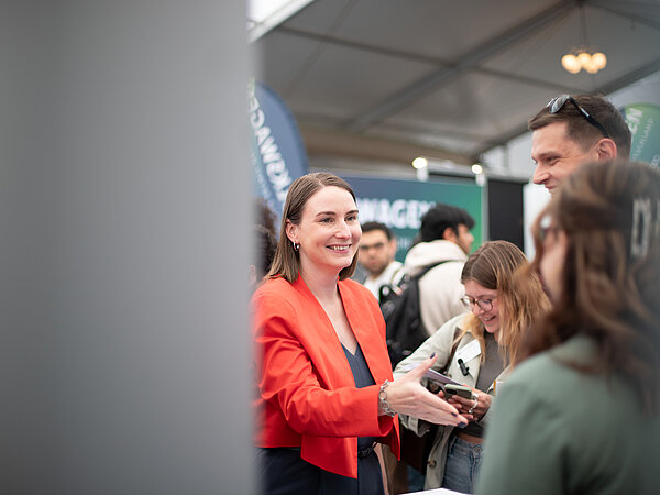 Frau im roten Blazer lächelt und spricht mit Besuchern auf einer Karrieremesse, umgeben von Messeständen und Gesprächsgruppen.