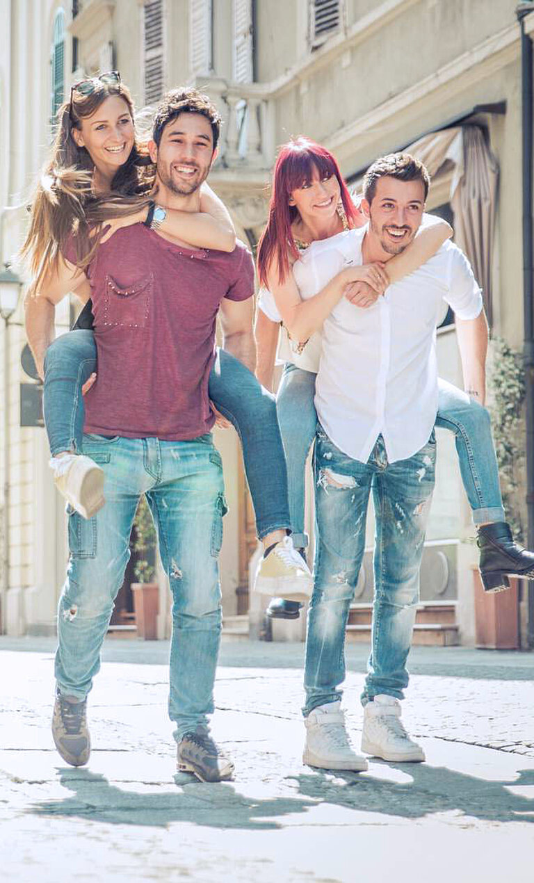 The image shows six friends walking through a city. Each man is carrying a woman on his back. They are all smiling and appear to be having a good time. Buildings and streetlights can be seen in the background, suggesting a bustling city.