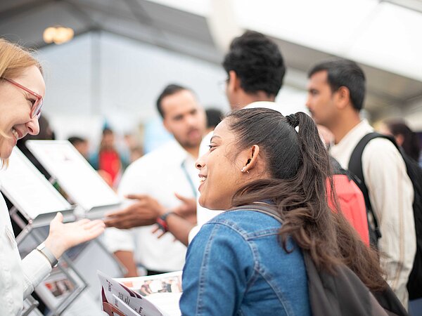 Zwei Frauen im Gespräch an einem Messestand in einem Zelt. Eine lächelt, während die andere gestikulierend etwas erklärt. Im Hintergrund sind weitere Besucher:innen und Informationsmaterialien sichtbar.