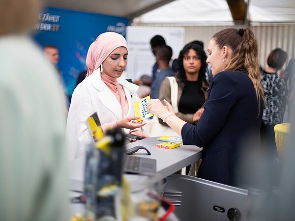 Zwei Frauen im Gespräch an einem Messestand, eine mit rosa Hijab und weißem Mantel präsentiert ein Produkt der Marke Kneipp, umgeben von weiteren Besucher:innen und Informationsmaterial.