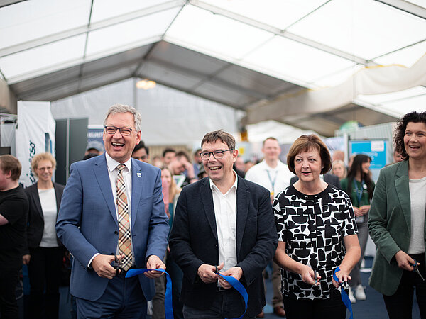Feierliche Eröffnung in einem großen Messezelt: Fünf Personen durchschneiden blaue Bänder vor einer Menschenmenge, umgeben von Messeständen und Ausstellungen