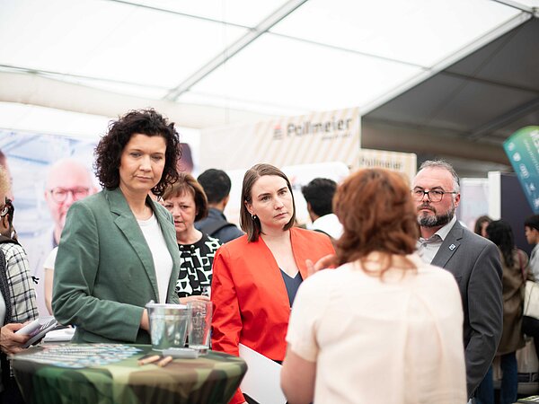 Personengruppe im Gespräch auf einer Messe in einem großen Zelt, im Vordergrund eine Person mit dem Rücken zur Kamera, gegenüber Zuhörer:innen in formeller Kleidung vor Informationsständen und Bannern.