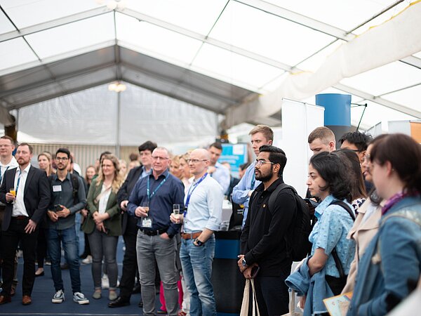 Große Gruppe von Personen steht in einem hellen Messezelt und hört aufmerksam einer Präsentation zu. Viele tragen Namensschilder und Lanyards, im Hintergrund sind Banner und Messestände sichtbar.