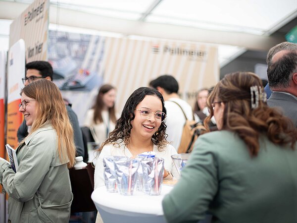 Personengruppe im Gespräch an einem Messestand mit silbernen Getränkepouches, im Hintergrund Banner mit der Aufschrift 'Pallmann' und weitere Informationsmaterialien.