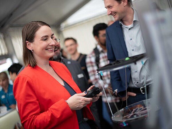 Frau im roten Blazer lächelt und hält einen Gamecontroller bei einer Technologie-Demonstration in einem Messezelt. Vor ihr ein Gerät in transparenter Hülle, daneben ein Mann im blauen Anzug, weitere Besucher:innen im Hintergrund.