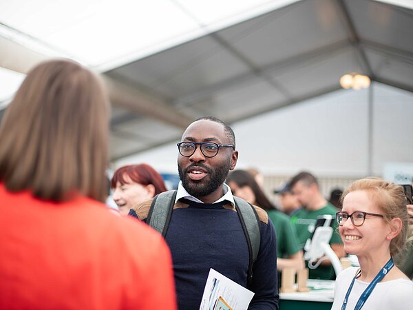 Personengruppe im Gespräch auf einer Karrieremesse in einem großen Zelt. Im Vordergrund ein Mann mit Brille und Rucksack spricht mit einer Frau im roten Blazer, weitere Besucher:innen sind im Hintergrund sichtbar.